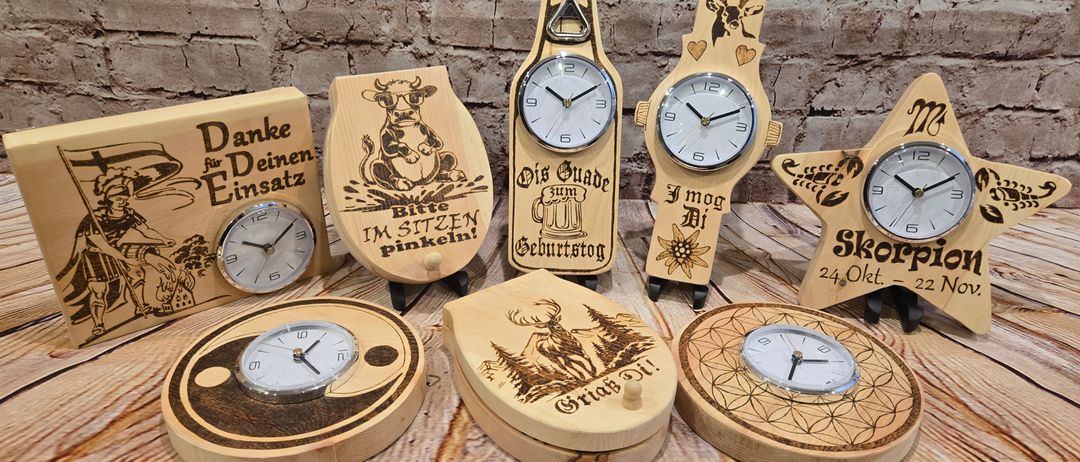 Mehrere Holzuhren mit einzigartigen Designs sind auf einer Holzoberfläche ausgestellt. Sie haben verschiedene Themen, einschließlich einer Kuh, eines Hirschs und eines Skorpions. Die Uhren haben unterschiedliche Designs und Texte auf den Zifferblättern.