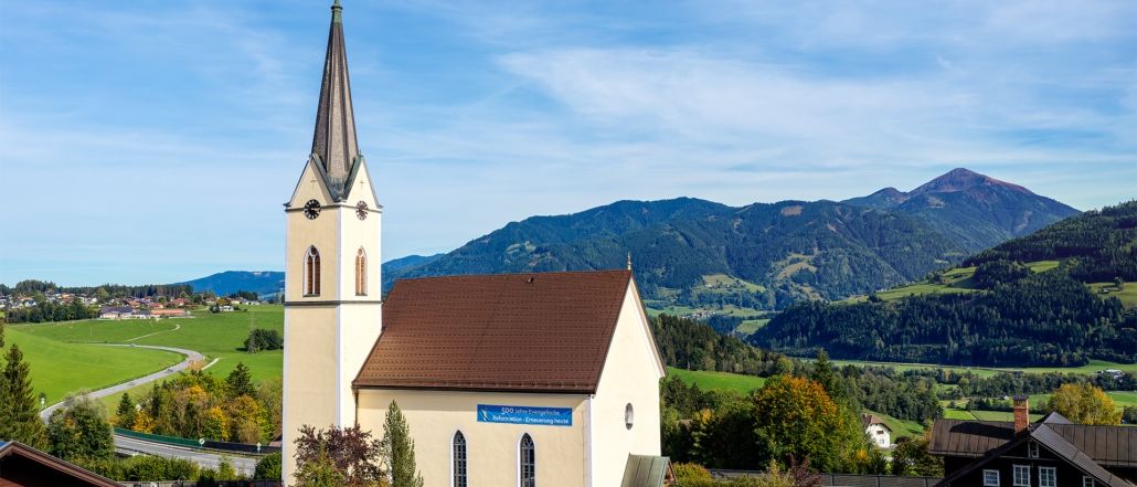 Eine Kirche mit braunem Dach und einem Turm, umgeben von einer malerischen Landschaft mit Bergen und Grünflächen, unter einem klaren blauen Himmel.