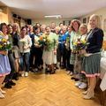 Eine Gruppe von Frauen steht in einem Raum mit Holzböden. Sie halten Blumensträuße in den Händen und tragen lässige bis halboffizielle Kleidung. Sie lächeln und scheinen für ein Gruppenfoto zu posieren.