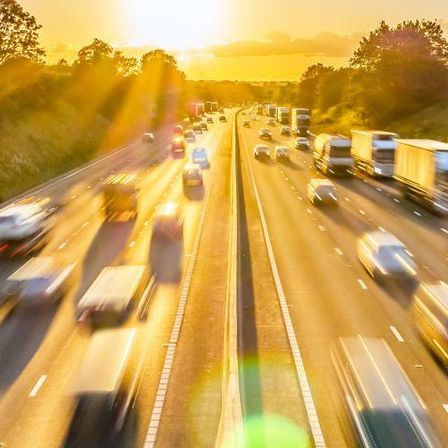 Eine vielbefahrene Autobahn mit Autos und Lastwagen in beide Richtungen, mit einem untergehenden Sonnenhintergrund.