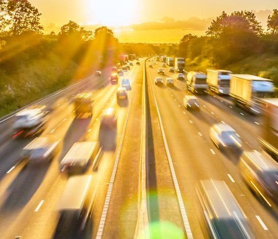 Eine vielbefahrene Autobahn mit Autos und Lastwagen in beide Richtungen, mit einem untergehenden Sonnenhintergrund.