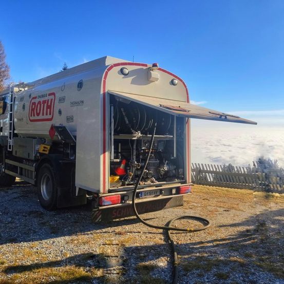 Ein weißer Wagen mit dem Wort 'Roth' auf der Seite, geparkt auf Kies mit offener Hintertür. Der Himmel ist klar, und im Hintergrund befindet sich ein Holzzaun.