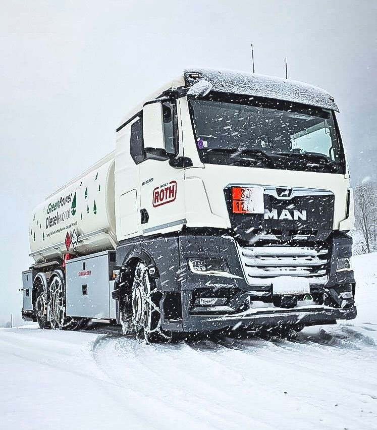 A white MAN truck with a tank for 'GreenPower' diesel-based fuel drives on a snowy road, equipped with snow chains and a license plate '12 34'.