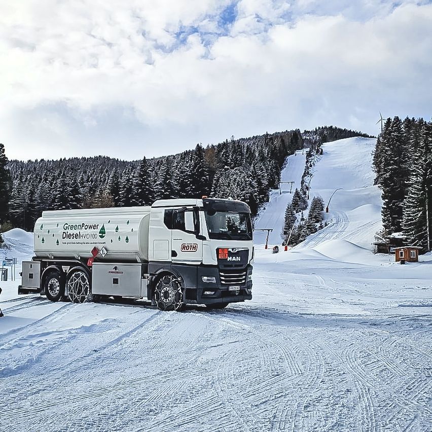 A white truck with GreenPower DieselHVO100 branding drives on a snowy road. Snow-covered trees and a ski slope are in the background.