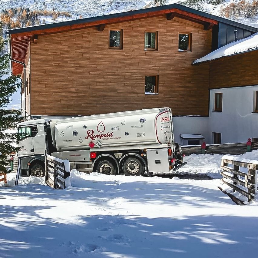 A white tanker truck labeled Rumbold is parked in front of a brown wooden house covered in snow.