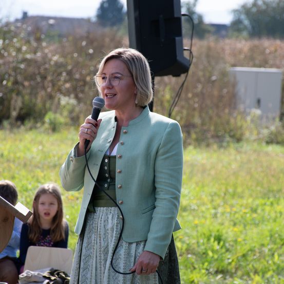 Eine Frau mit Brille spricht in ein Mikrofon auf einem Feld. Hinter ihr steht ein Lautsprecher und einige Leute. Ein Kind sitzt und schaut sie an.