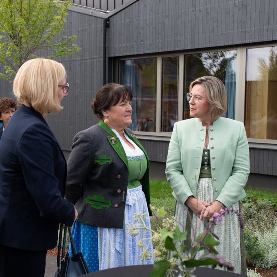Drei Frauen, zwei in traditioneller Kleidung, unterhalten sich in der Nähe eines Gebäudes mit Glasfenstern, einem Baum und einem Rasenbereich.