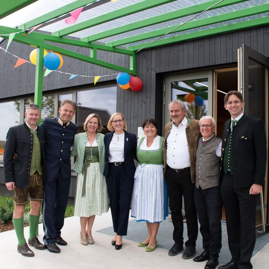 Eine Gruppe von Personen, darunter Männer und Frauen, in traditioneller Kleidung, stehen vor einem Gebäude mit Dekorationen und lächeln für ein Foto.