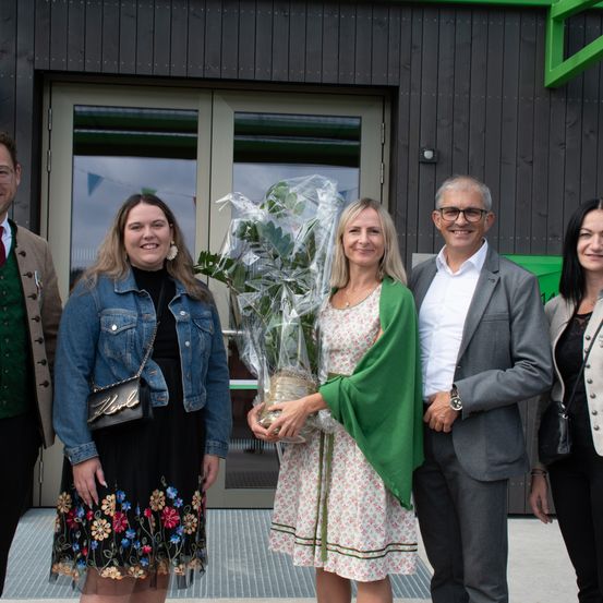 Fünf Menschen stehen vor einem modernen Gebäude. Eine Frau hält eine Pflanze. Sie alle sehen glücklich aus.