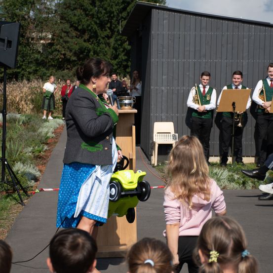 Eine Frau steht an einem Rednerpult und spricht eine Gruppe von Kindern an. Dahinter bereitet sich eine Gruppe von Musikern in grünen Westen und schwarzen Hosen auf eine Aufführung vor.