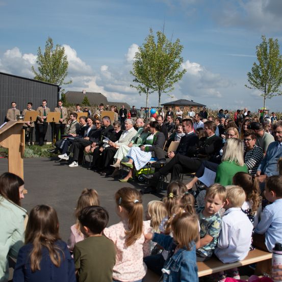 Eine große Menschenmenge, einschließlich Erwachsener und Kinder, sitzt auf Bänken und Stühlen und beobachtet eine Veranstaltung im Freien. In der Vorderreihe ist ein Holzpodium zu sehen, und eine Person steht daneben. Dahinter stehen mehrere Personen, möglicherweise Mitglieder einer Band, mit Instrumenten auf Ständern. Im Hintergrund sind Bäume und ein Gebäude zu sehen.