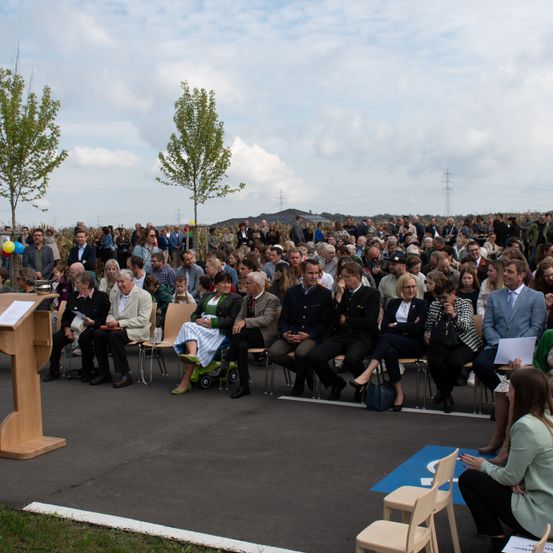 Eine Menschenmenge in Anzügen und Kleidern sitzt auf Stühlen in einem Parkplatz und nimmt an einer Veranstaltung teil. Einige halten Papiere und ein Podium mit einem Buch steht links. Bäume und Strommasten sind im Hintergrund.