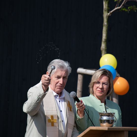 Ein älterer Mann und eine Frau stehen an einem Podium mit Luftballons. Der Mann hält eine Flasche und scheint Flüssigkeit zu versprühen, während die Frau ein Mikrofon hält.