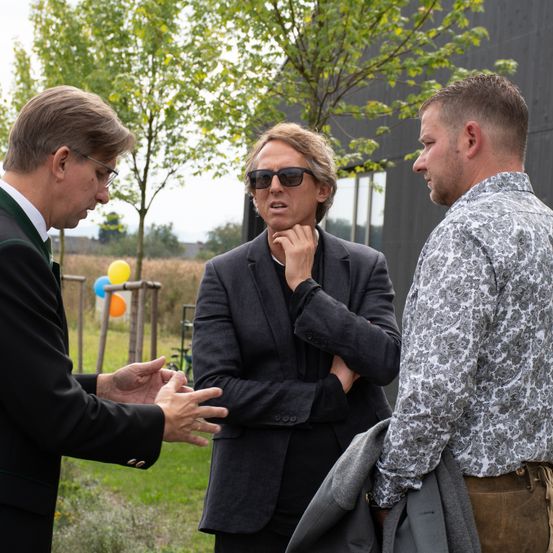 Drei Männer unterhalten sich draußen in der Nähe eines Gebäudes. Einer trägt einen Anzug, ein anderer ein gemustertes Hemd und der dritte einen grünen Weste. Im Hintergrund sind Bäume und Luftballons zu sehen.