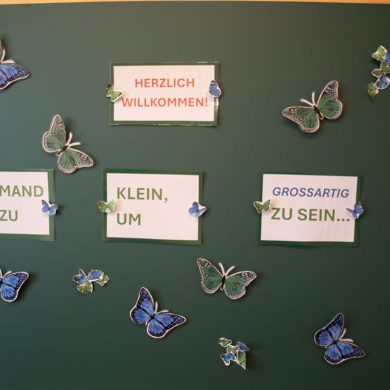 Eine grüne Tafel mit Schmetterlingen und Papierausschnitten, die 'Klein, um' und 'Grossartig zu sein' sagen, mit 'Herzlich willkommen!' oben. Viele blaue und grüne Schmetterlinge sind an der Tafel befestigt.