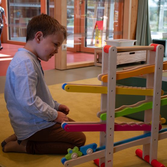 Ein junger Junge sitzt auf dem Boden und spielt mit einem farbenfrohen Holzspielzeug. Er lächelt und scheint sich zu amüsieren. Das Spielzeug hat verschiedene Bahnen und Kugeln. Hinter ihm befindet sich eine Glastür mit Blick auf das Freie.