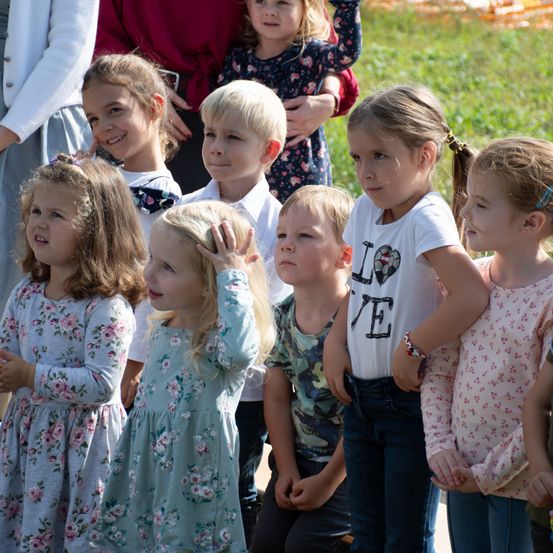 Eine Gruppe von Kindern, einige stehend und andere kauernd, schauen in die gleiche Richtung. Ein Mädchen berührt seine Stirn mit der Hand. Hinter ihnen stehen Erwachsene, darunter eine Frau in einem weißen Shirt.