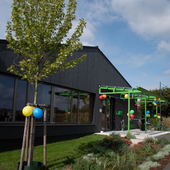 Ein modernes Gebäude mit Glasfenstern, einer schwarzen Außenfassade und einer grünen Pergola mit bunten Luftballons. Vor dem Gebäude steht ein Baum mit Luftballons.