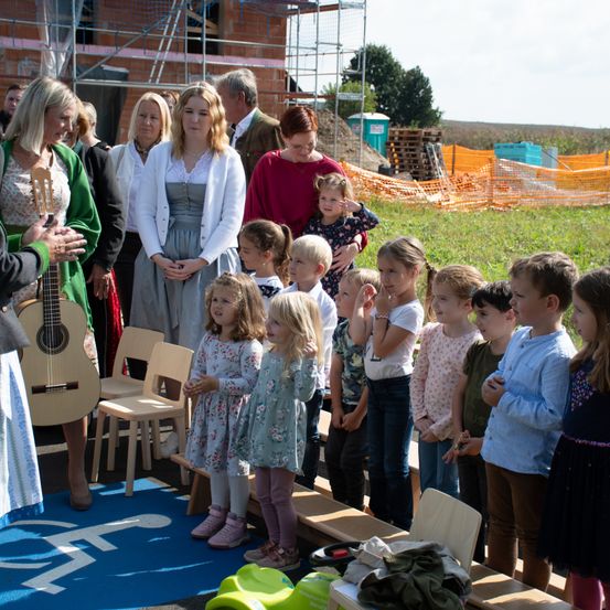 Eine Gruppe von Erwachsenen und Kindern steht im Freien, wobei eine Frau eine Gitarre hält. Hinter ihnen befinden sich ein Bauplatz und orangefarbene Zäune. Die Kinder schauen etwas vor sich an. Der Boden ist mit einem blauen Teppich bedeckt, auf dem ein weißes Schild liegt.