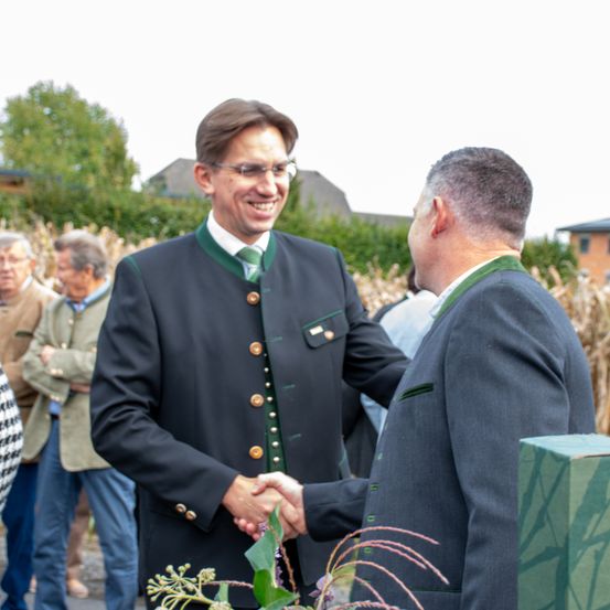 Zwei Männer in Anzügen schütteln sich draußen die Hände und lächeln. Einer trägt eine grüne Krawatte und Knöpfe. Verschwommene Menschen und Pflanzen im Hintergrund.