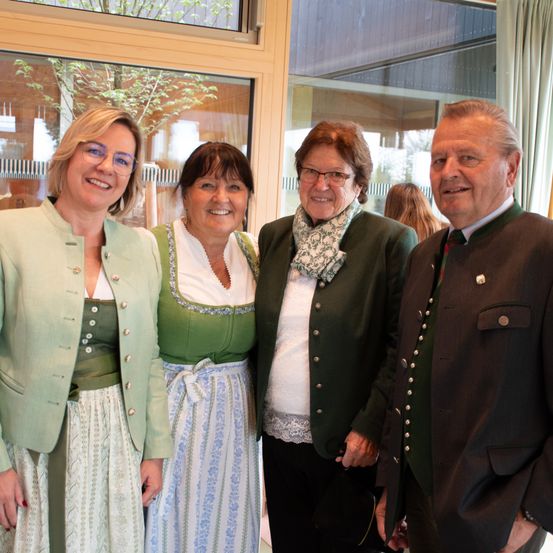 Vier Personen, gekleidet in traditioneller Kleidung, stehen gemeinsam in einem Raum. Zwei Frauen tragen grüne und weiße Kleider mit Schals, während die anderen beiden in formellen Anzügen sind. Sie scheinen zu lächeln und posieren für ein Foto.
