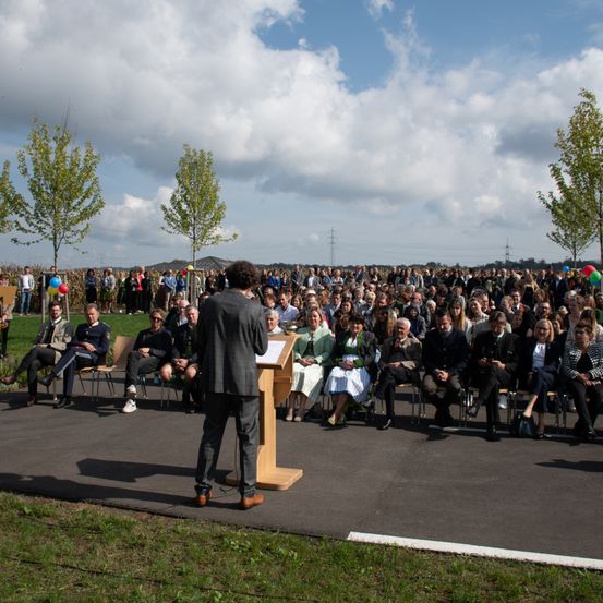 Ein Mann steht an einem Podium und spricht eine große Menschenmenge an. Das Publikum sitzt auf Stühlen, einige stehen. Bunte Luftballons sind vorhanden. Die Umgebung umfasst Bäume und einen klaren Himmel.
