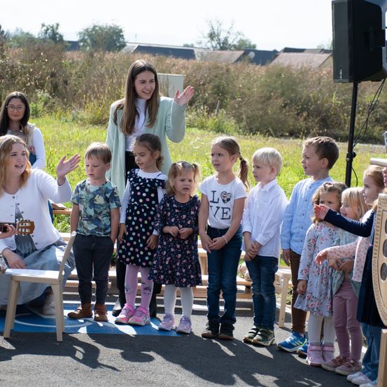 Eine Gruppe von Kindern und Erwachsenen steht in einer Reihe, lächelt und posiert für ein Foto. Eine Frau spielt links eine Gitarre. Die Kinder tragen verschiedene Kleidungsstücke und Schuhe. Der Hintergrund ist eine grasbewachsene Fläche mit einem Haus und Bäumen.