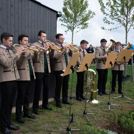 Eine Brassband von Männern in gleichen Uniformen steht auf einem Rasenfeld und spielt Trompeten und Tuben. Dahinter befindet sich ein schwarzes Gebäude mit Bäumen im Hintergrund.