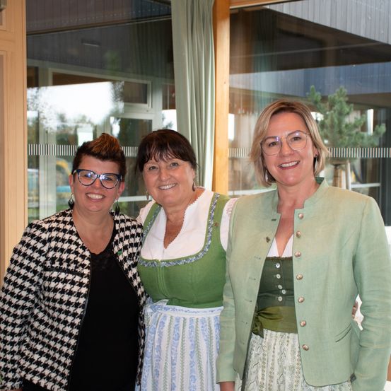 Drei Frauen stehen eng beieinander und lächeln für ein Foto vor einem Glasfenster. Die linke Frau trägt eine Brille und eine schwarz-weiß karierte Jacke. Die mittlere Frau trägt ein grünes und weißes traditionelles Kleid. Die rechte Frau trägt eine grüne Jacke und ein grünes und weißes Kleid.