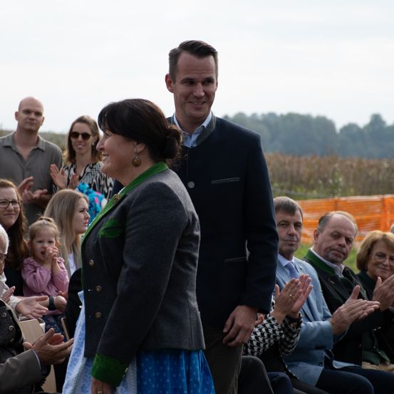 Eine Gruppe von Menschen ist im Freien versammelt. Ein Mann und eine Frau stehen vor anderen, die klatschen und lächeln. Sie befinden sich auf einem Feld mit Bäumen und orangefarbenen Barrieren.