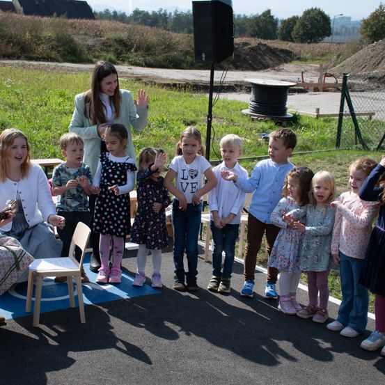 Eine Gruppe junger Kinder, angeleitet von einer Frau, steht auf einer Straße, wahrscheinlich für eine Schulveranstaltung. Ein Lautsprecher steht hinter ihnen, und ein Stuhl ist in der Nähe.