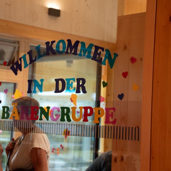 Eine Frau steht neben einer Glastür mit einem Schild, das 'Willkommen in der Barengruppe' sagt. Das Schild ist mit farbenfrohen Herzen verziert. Ein Mann ist teilweise auf der anderen Seite sichtbar.