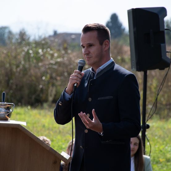 Ein Mann spricht in ein Mikrofon an einem Rednerpult, mit einem Lautsprecher dahinter, während Leute zuschauen. Im Hintergrund befindet sich ein grasbewachsenes Feld.