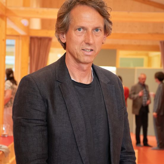 Ein Mann in einem dunklen Anzug steht drinnen und schaut direkt in die Kamera. Hinter ihm unterhalten sich zwei Personen. Der Hintergrund besteht aus Holzbalken und einem roten Boden.