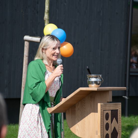 Eine Frau steht hinter einem Rednerpult, hält ein Mikrofon und lächelt. Hinter ihr befindet sich ein Holzpfosten mit bunten Luftballons. Ein silberner Becher steht auf dem Pult. Ein schwarzer Hintergrund ist im Hintergrund zu sehen.