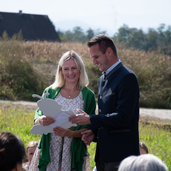Ein Mann und eine Frau stehen Hand in Hand in einem Feld, lächeln und schauen auf ein Stück Papier. Im Hintergrund befinden sich Kinder und ein Haus.