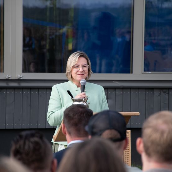 Eine Frau in einem mintgrünen Anzug steht an einem Rednerpult, hält ein Mikrofon in der Hand und spricht zu einer Gruppe von Menschen vor ihr. Das Publikum ist teilweise verschwommen. Die Umgebung scheint sich im Freien abzuspielen.