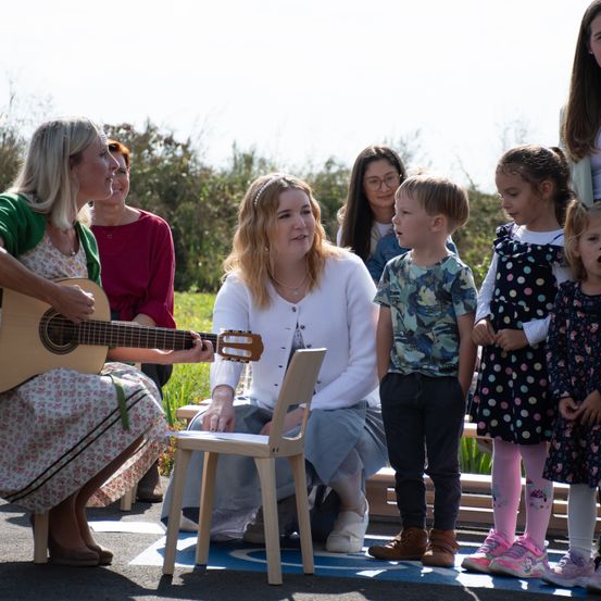 Eine Frau mit einer Gitarre sitzt auf einem Stuhl, umgeben von Kindern, darunter ein Junge im Camouflage-Shirt und ein Mädchen im Punktekleid. Andere Erwachsene sind ebenfalls anwesend, einige stehen hinter ihnen.