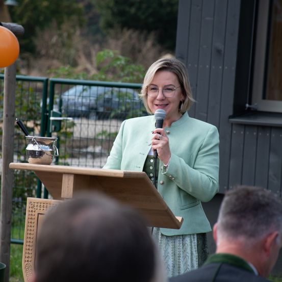 Eine Frau in einer hellgrünen Jacke steht an einem Rednerpult und hält ein Mikrofon. Sie scheint zu einem kleinen Publikum zu sprechen. Hinter ihr befindet sich ein Holzzaun, ein kleiner oranger Ballon und eine silberne Kanne auf dem Podest.