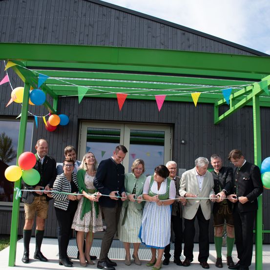 Eine Gruppe von Menschen steht vor einem Gebäude, schneidet ein Band und hat Luftballons und Wimpel im Hintergrund.