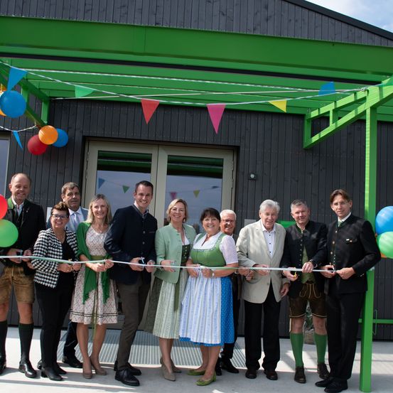 Eine Gruppe von Menschen steht vor einem Gebäude. Sie schneiden ein Band und lächeln. Es gibt Luftballons und Wimpelgirlanden als Dekoration. Das Gebäude hat Glasfenster und eine Tür.