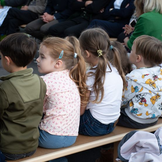 Vier Kinder sitzen auf einer Bank und schauen aufmerksam etwas an. Das Mädchen in der Mitte hat rosa Haare und eine blaue Haarnadel. Rechts trägt ein Junge ein Mickey-Mouse-Hoodie.