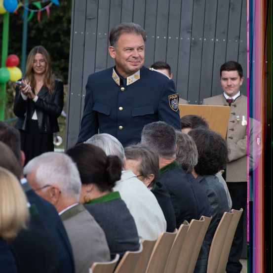 Ein Mann in Uniform steht vor einer sitzenden Menge. Eine Frau macht ein Foto mit einer Kamera. Der Anlass scheint feierlich zu sein, mit Luftballons und einem Podium im Hintergrund.