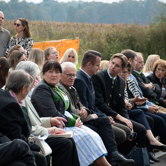 Eine Gruppe von Menschen sitzt auf einer Bank bei einer Veranstaltung im Freien. Einige tragen Brillen und formelle Kleidung. Hinter ihnen sind ein Feld mit hohem Gras und orangefarbene Barrieren zu sehen.