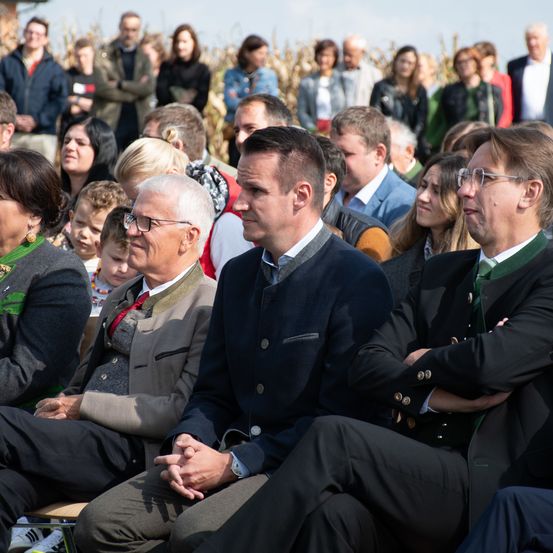 Eine große Menschenmenge ist in einem Maisfeld versammelt. Einige sitzen, andere stehen. Einige von ihnen tragen Brillen und einige tragen Krawatten.