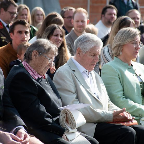 Eine Gruppe von Menschen sitzt und steht in einer Reihe. Sie scheinen an einer formellen Veranstaltung teilzunehmen. Eine ältere Frau mit Brille und einer karierten Schürze sitzt neben einem älteren Mann. Eine Frau in einem grünen Oberteil sitzt neben ihnen. Dahinter sitzen eine Gruppe von Menschen auf Stühlen.