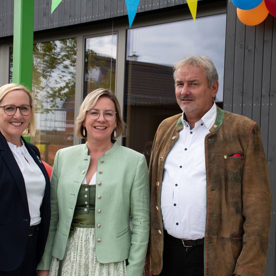 Drei Personen stehen vor einem Gebäude. Die Frau links trägt eine Brille und einen marineblauen Blazer. Die mittlere Frau hat eine grüne Jacke und einen weißen Rock. Der Mann rechts trägt einen braunen Ledermantel. Hinter ihnen befinden sich Luftballons und ein Fenster.