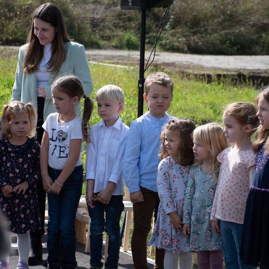 Eine Frau und mehrere Kinder stehen in einer Reihe, möglicherweise auf einem Feld. Die Frau trägt eine Jacke und scheint zu lächeln. Die Kinder tragen verschiedene Kleidungsstücke und einige schauen die Frau an.
