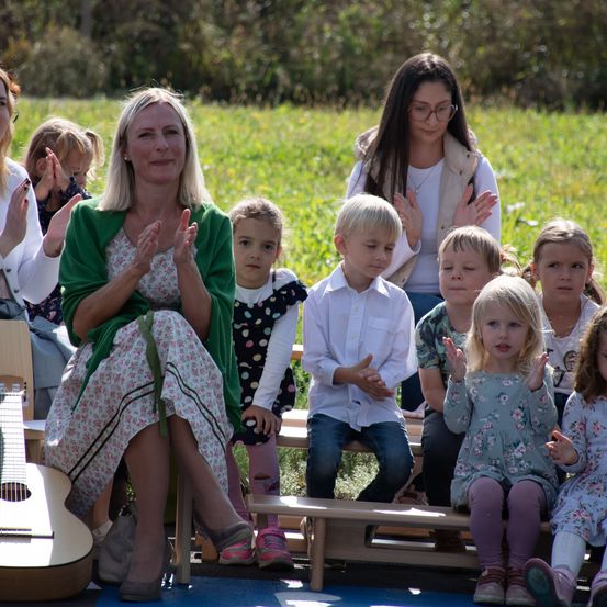 Eine Gruppe von Kindern und Erwachsenen sitzt auf einer grasbewachsenen Wiese und klatscht in die Hände. Eine Frau in Grün und eine Frau in Weiß stehen in der Nähe, und eine kleine Gitarre liegt auf dem Boden.