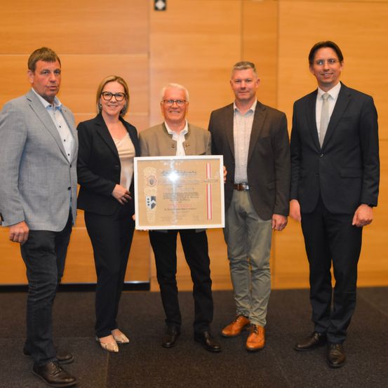 Fünf Männer in Anzügen posieren für ein Foto und halten eine Urkunde. Der Mann in der Mitte hält eine Urkunde. Sie stehen vor einer Holzwand.